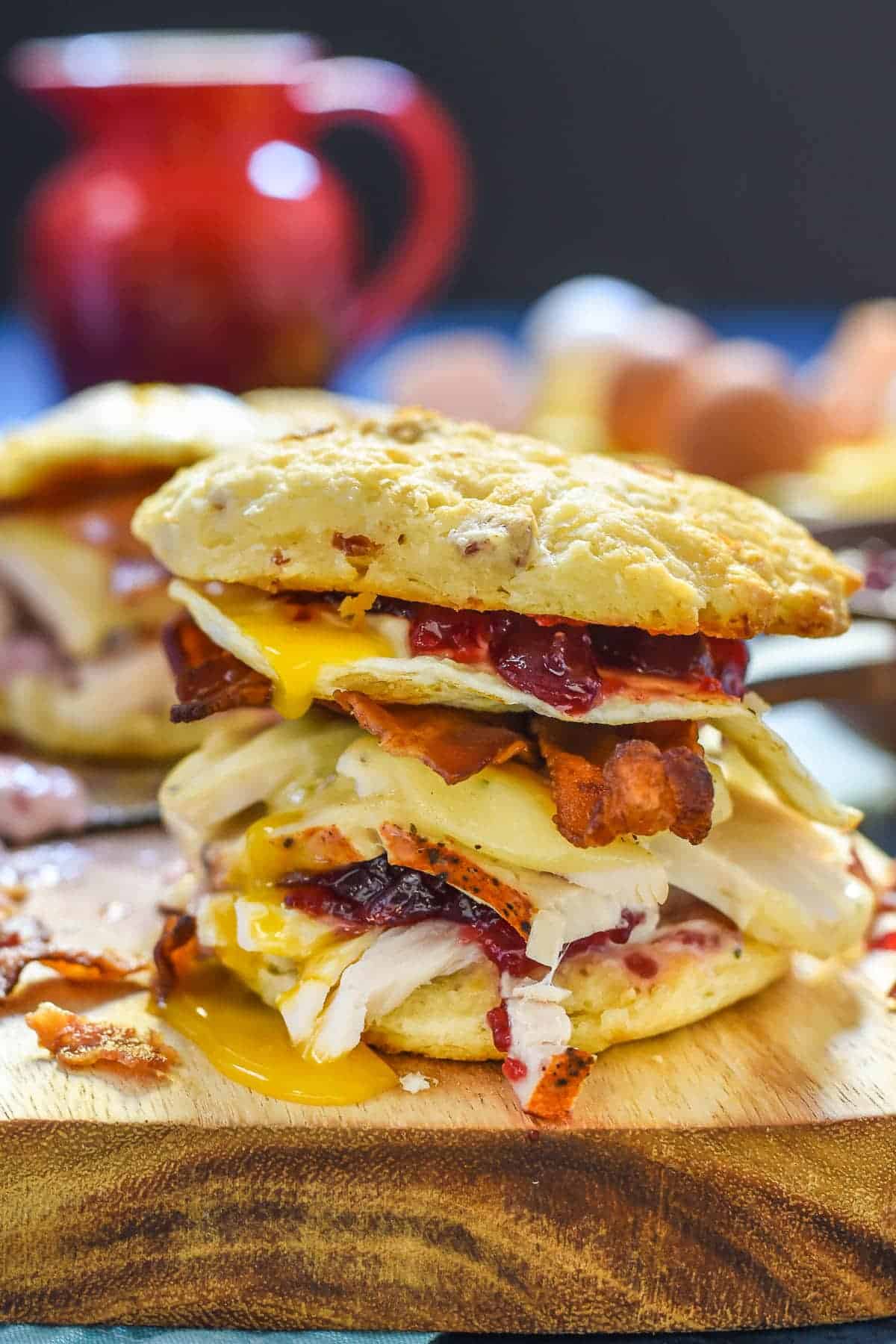 A towering sandwich with layers of turkey, bacon, melted cheese, cranberry sauce, and a runny egg between two golden, crispy buns. It's set on a wooden board with a blurred red mug and eggs in the background.