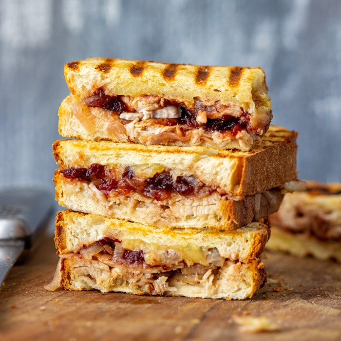 A stack of grilled turkey and cranberry sandwiches on toasted bread sits on a wooden board. The sandwiches are cut in halves, revealing juicy turkey and vibrant cranberry sauce inside. A blurred gray background completes the scene.