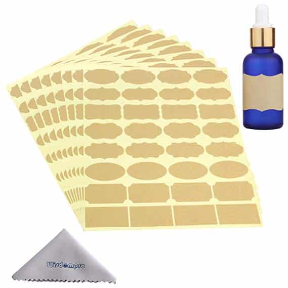 Sheets of gold adhesive labels in various shapes accompany a blue glass dropper bottle adorned with a matching gold label—an elegant presentation ideal for gifts in a jar. A small gray microfiber cloth is also included.