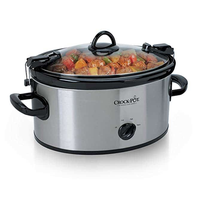 A silver Crock-Pot slow cooker with a black handle and a glass lid, filled with a variety of colorful vegetables and chunks of meat. The cooker is set on a white surface.