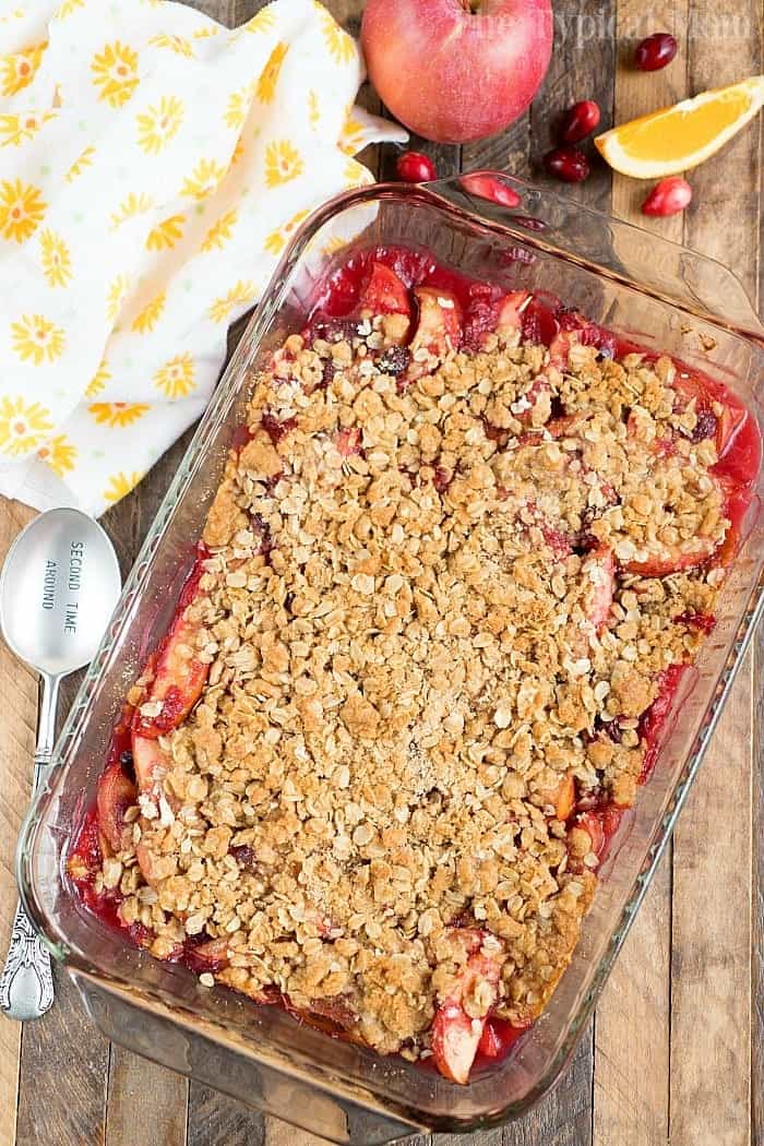 A glass baking dish filled with apple cranberry crisp topped with a golden oat and brown sugar crumble. A spoon labeled "Scoop Me" rests beside it, with a peach and cranberries nearby. A yellow floral-patterned cloth is partially visible.