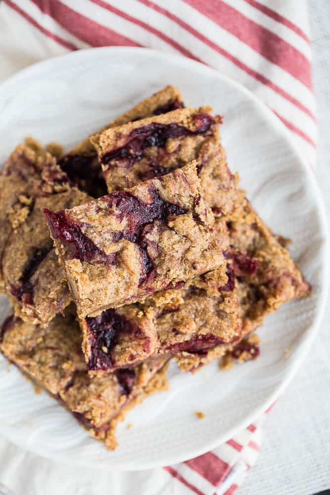 A plate of rustic, crumbly bar cookies with swirls of red fruit topping sits on a striped red and white cloth. The bars have a textured, homemade appearance.