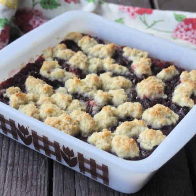 A freshly baked cobbler in a square dish on a wooden surface. The dessert has a golden, crumbly topping with visible berry filling underneath. The dish features a decorative brown pattern on its sides. A floral-patterned cloth is partially visible.