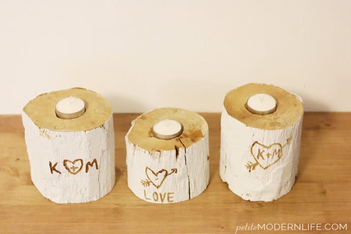Three rustic candle holders made from tree trunks with white painted bark, each featuring carved heart designs. The carvings read "K + M" and "LOVE" with an arrow through a heart. A small white candle sits on top of each holder.