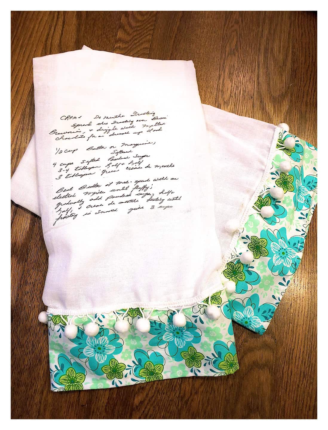 A white linen towel with hand-written text lies on a wooden surface. It features a colorful floral trim with shades of green and blue, adorned with small white pom-poms.