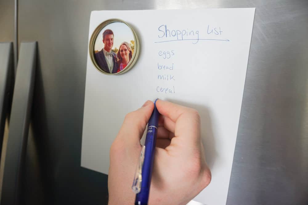A hand writes a shopping list on a white sheet of paper attached to a refrigerator with a round magnet featuring a photo of a couple. The list includes eggs, bread, milk, and cereal.