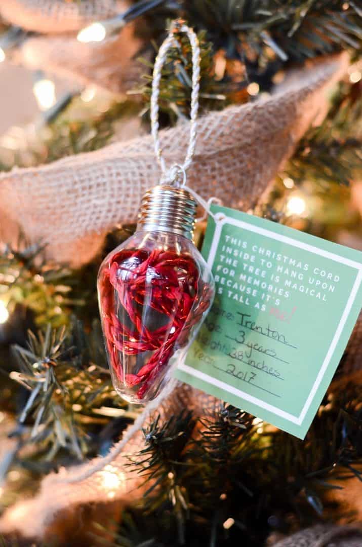 A Christmas tree ornament shaped like a lightbulb filled with red tinsel hangs on a tree. Attached to the ornament is a green tag with handwritten text, surrounded by burlap garland and white lights.