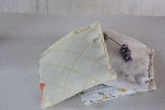 Three small, handmade fabric sachets are stacked on a wooden surface. The top sachet has a geometric pattern, and a sprig of lavender lies on it. The other sachets have subtle pastel prints. The background is light and textured.