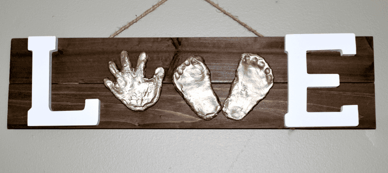 A wooden wall hanging spells "LOVE" with white wooden letters "L" and "E." The "O" and "V" are represented by a gold-painted handprint and pair of footprints.
