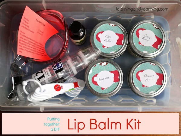 Top-down view of a DIY lip balm kit in a plastic container. Includes jars labeled Shea Butter, Beeswax, Cocoa Butter, and Coconut Oil, a clear pipette, red measuring spoons, a small bottle, and a red instruction sheet. Text reads "Lip Balm Kit.