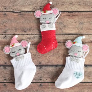 Three felt mice wearing Santa hats snuggled inside stockings on a wooden surface. Two mice are in white stockings with "2018" on one, and the third is in a red stocking. Each mouse has gray fur and rosy cheeks.