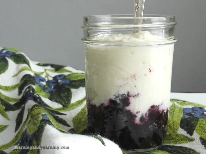A jar of homemade yogurt with a layer of dark fruit compote at the bottom, placed on a floral-patterned cloth. A spoon is inserted into the yogurt.