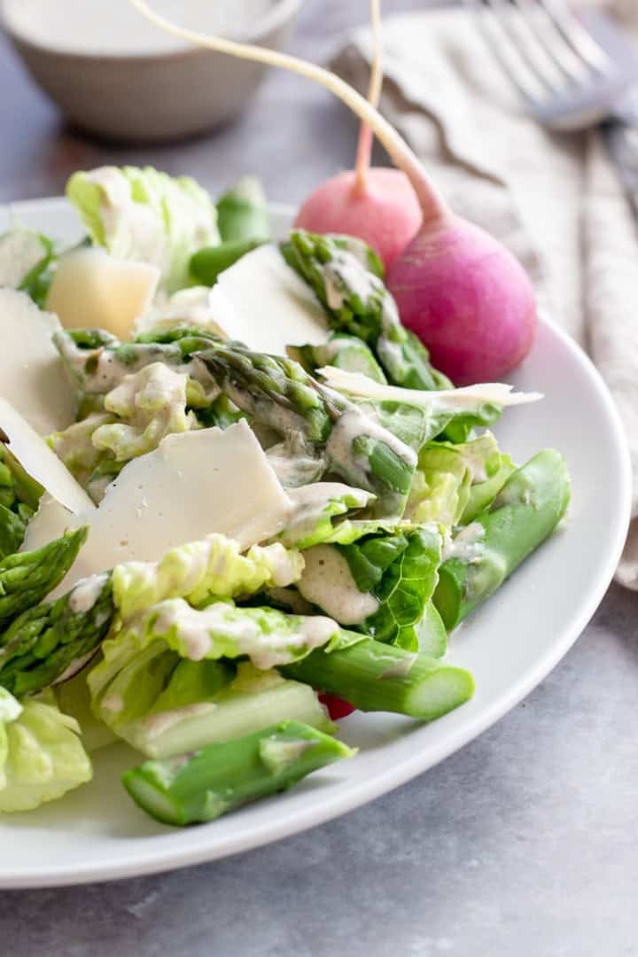 A fresh spring salad featuring romaine lettuce, asparagus, radish, and shaved cheese is drizzled with creamy dressing on a white plate. A fork and napkin are in the background.