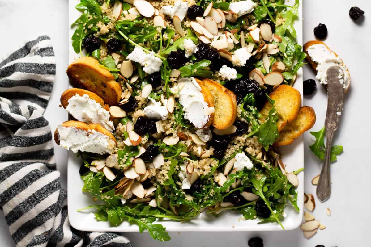 A fresh spring salad with arugula, goat cheese, toasted almonds, and dried cherries is elegantly presented on a white rectangular platter. Slices of toasted baguette are artfully scattered on top. A striped napkin and a knife rest beside the platter, perfect for enjoying this seasonal delight.