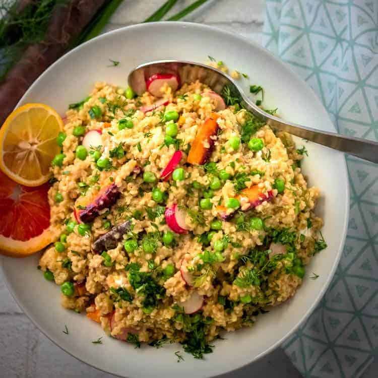 A delightful spring salad featuring couscous, green peas, and sliced radishes, all tossed with fresh herbs. Garnished with lemon and orange slices on the side, this vibrant dish is perfect for exploring new salad recipes. A spoon rests invitingly on the bowl.