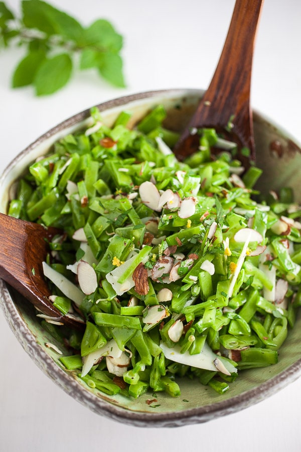 A fresh spring salad in a bowl featuring chopped green beans, sliced almonds, and herbs, with two wooden serving spoons. A sprig of fresh herbs is in the background on the white surface. Perfect for those exploring spring salad recipes!