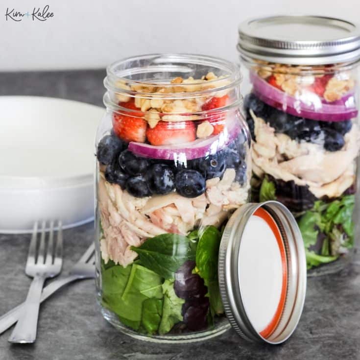 Two mason jars filled with vibrant spring salad recipes, featuring layers of spinach, chicken, blueberries, red onions, strawberries, and walnuts. One jar is open to reveal its colorful contents. Plates and forks await on a gray surface.