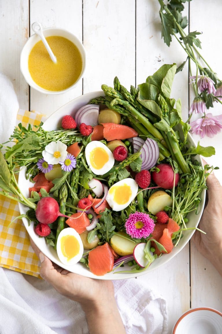 A vibrant spring salad recipe featuring a bowl of boiled eggs, radishes, asparagus, red onions, raspberries, and flowers. Bursting with assorted greens and colorful vegetables, it includes a small bowl of yellow dressing on the side for a bright, fresh presentation on a white table.