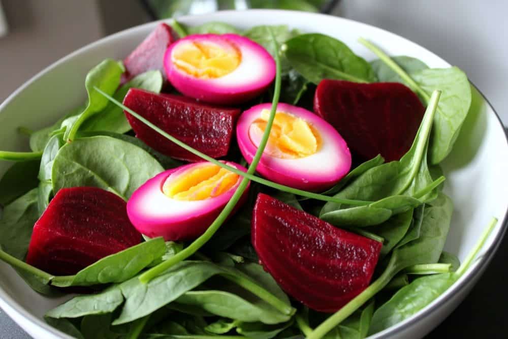 A bowl of fresh spinach, perfect for spring salad recipes, topped with slices of pickled eggs showcasing vibrant pink layers and yellow yolks, alongside pieces of red beetroot.