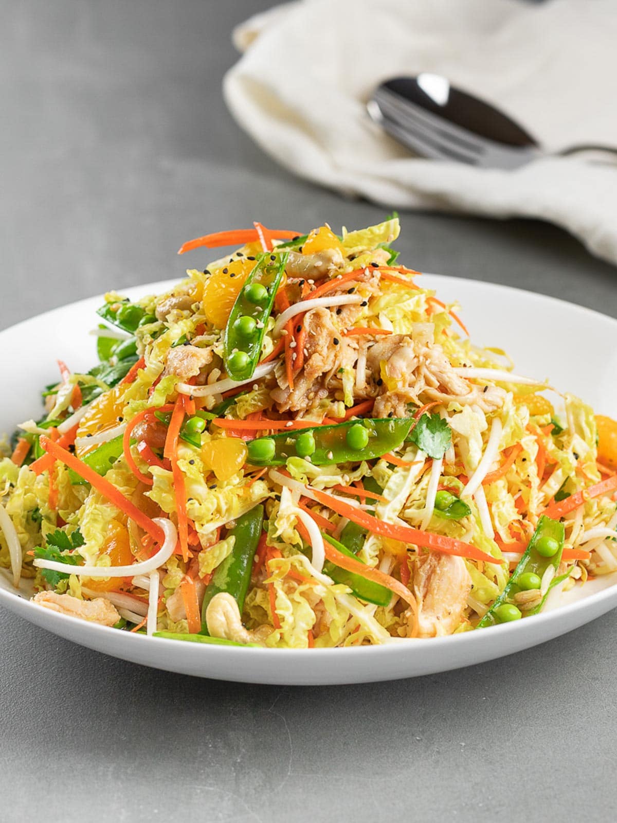 A fresh spring salad on a white plate, featuring shredded cabbage, carrots, snow peas, bean sprouts, cooked chicken, and mandarin orange slices. The dish is garnished with cilantro and black pepper. A napkin and cutlery are in the background; a delightful addition to your spring salad recipes.