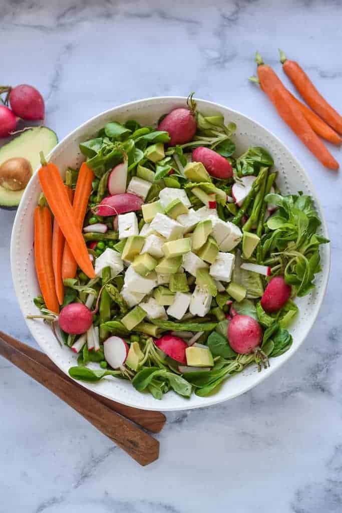 A fresh spring salad recipe featuring leafy greens, sliced radishes, cubed avocado, and chunks of feta cheese. It's garnished with whole radishes, carrot sticks, and avocado halves on a marble surface. Wooden salad tongs are nearby for serving this vibrant dish.