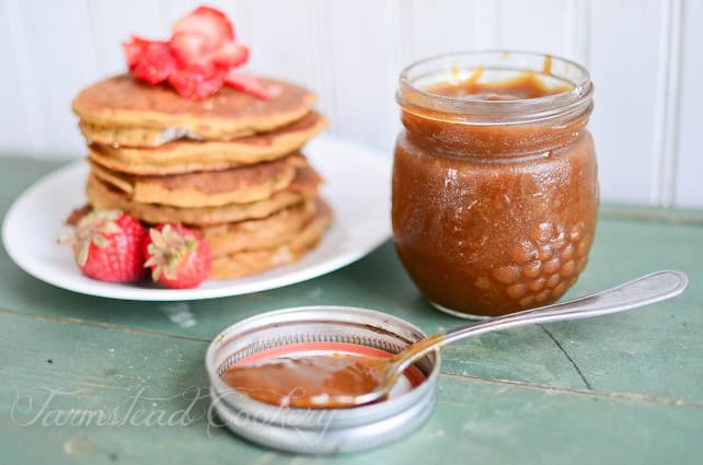 A stack of pancakes topped with sliced strawberries sits next to a jar of apple butter, surrounded by a green table—a delightful spread that inspires creative breakfast ideas. Consider adding ways to preserve and use rhubarb as a tangy twist. Two whole strawberries rest on the plate against a wooden backdrop.