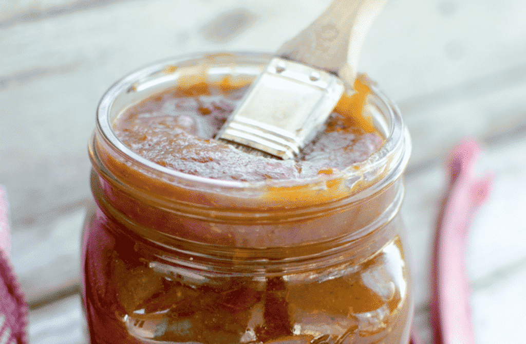 A jar filled with reddish-brown barbecue sauce sits invitingly, with a small wooden brush resting on top, partially dipped into the tangy mix. The rustic wood background and hints of red cloth are complemented by fresh rhubarb stalks, showcasing creative ways to preserve and use rhubarb in culinary delights.