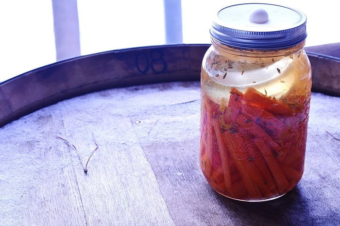 A jar filled with fermented carrot sticks, partially submerged in pickling liquid, sits on a wooden surface. Sunlight gently illuminates the jar, highlighting details of the contents. The background is softly blurred, providing a calm and rustic atmosphere.
