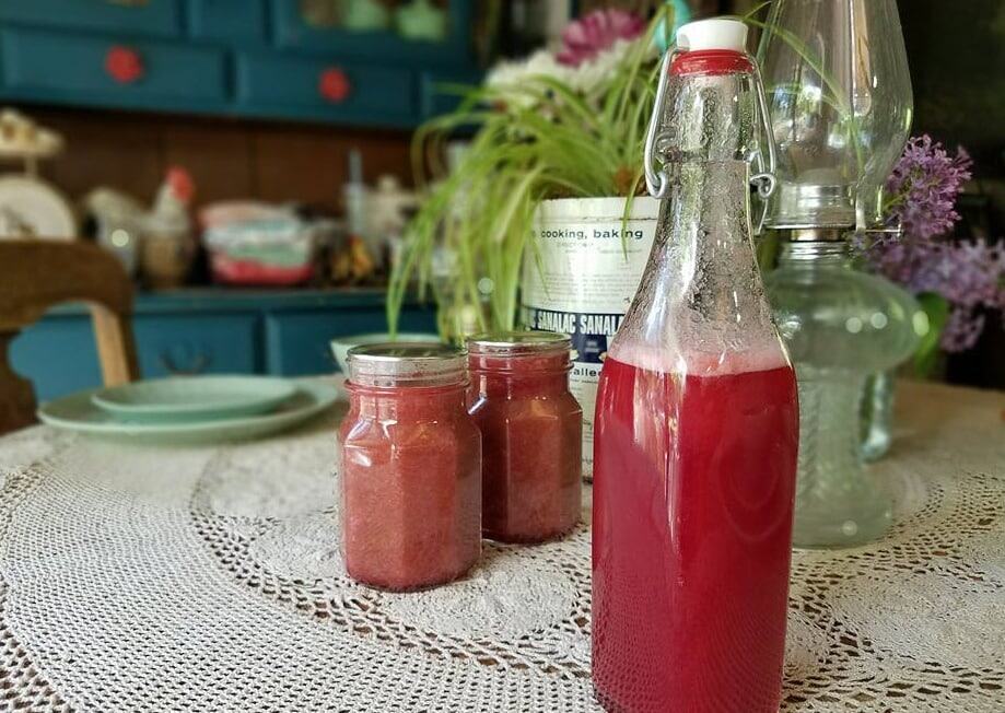 A glass bottle and two jars filled with red liquid, showcasing creative ways to preserve and use rhubarb, sit on a lace tablecloth. In the background, a kitchen with a blue cabinet, plates, and an oil lamp creates a charming scene. Some lavender flowers are also visible.