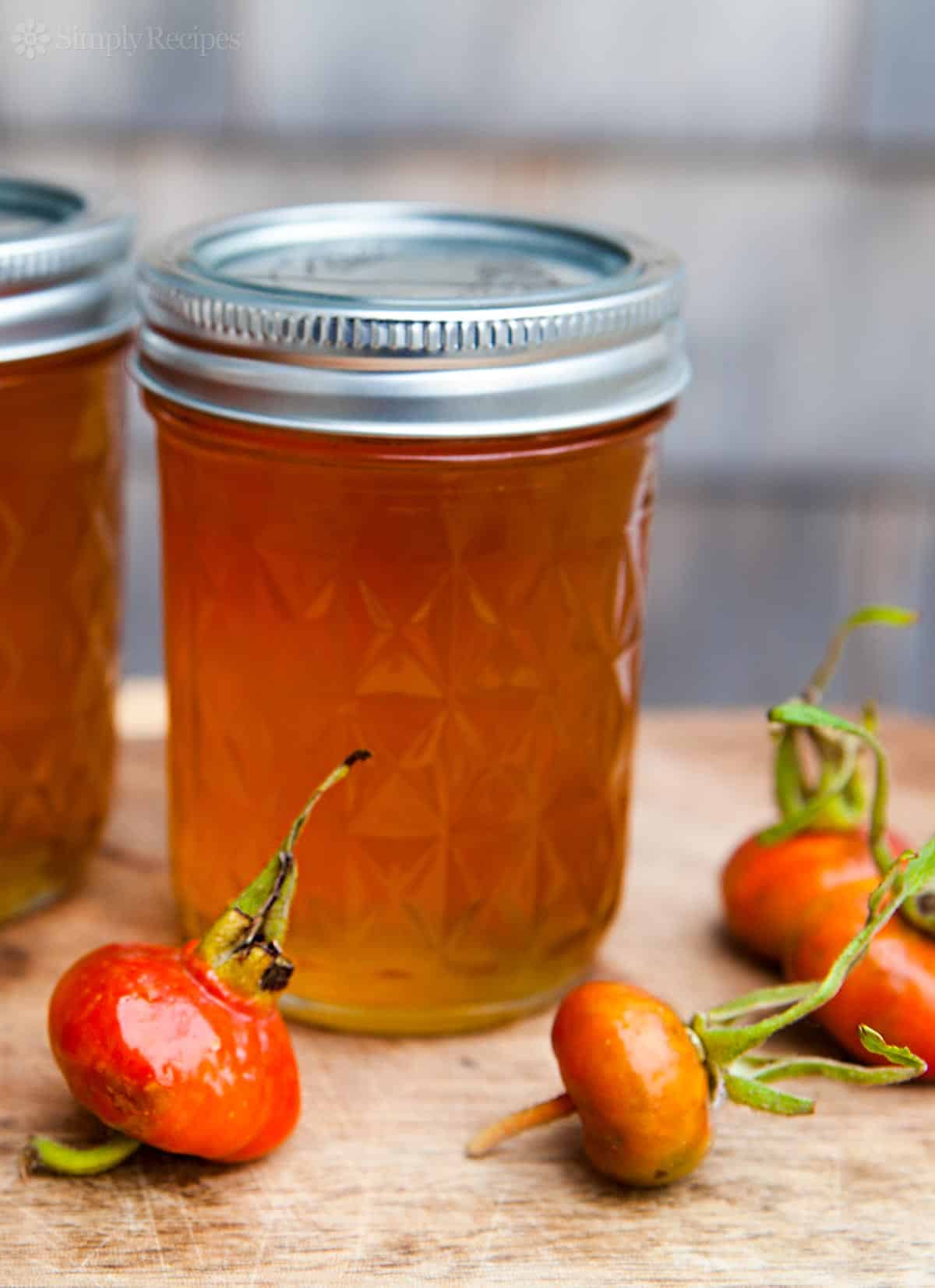 A jar of golden rose hip jelly with a diamond-patterned lid sits on a wooden surface, inviting you to explore the art of making flower jellies. Fresh rose hips are placed around the jar.