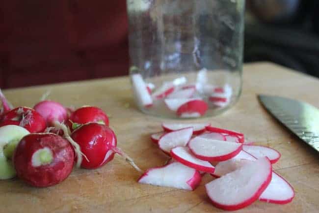 Fresh radishes, some whole and some sliced, are placed on a wooden cutting board. A knife is nearby, and a glass jar partially filled with fermented radish slices is in the background.