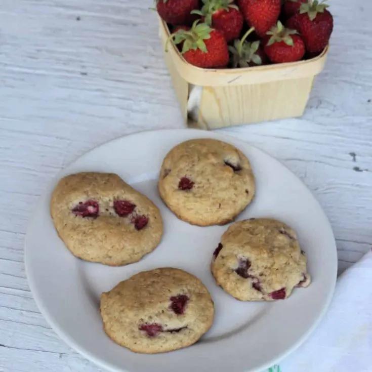 4 strawberry fruit cookies on a white plate sitting in front of a basket of whole strawberries.