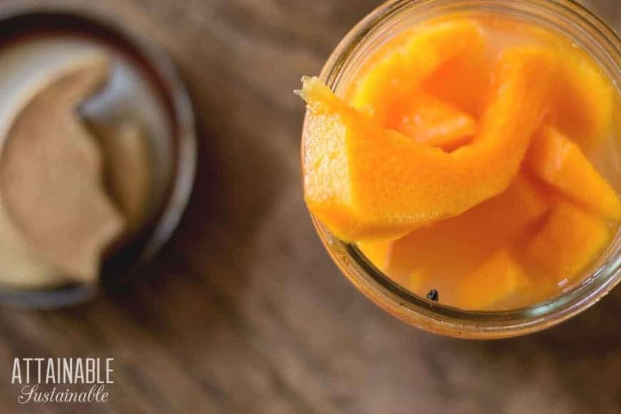 A glass jar filled with bright orange peach slices and liquid, viewed from above. On the wooden surface, a small brown dish with a crescent shape is in the background. The words "Attainable Sustainable" are printed in the lower left corner, evoking thoughts of fermented vegetables and wholesome living.