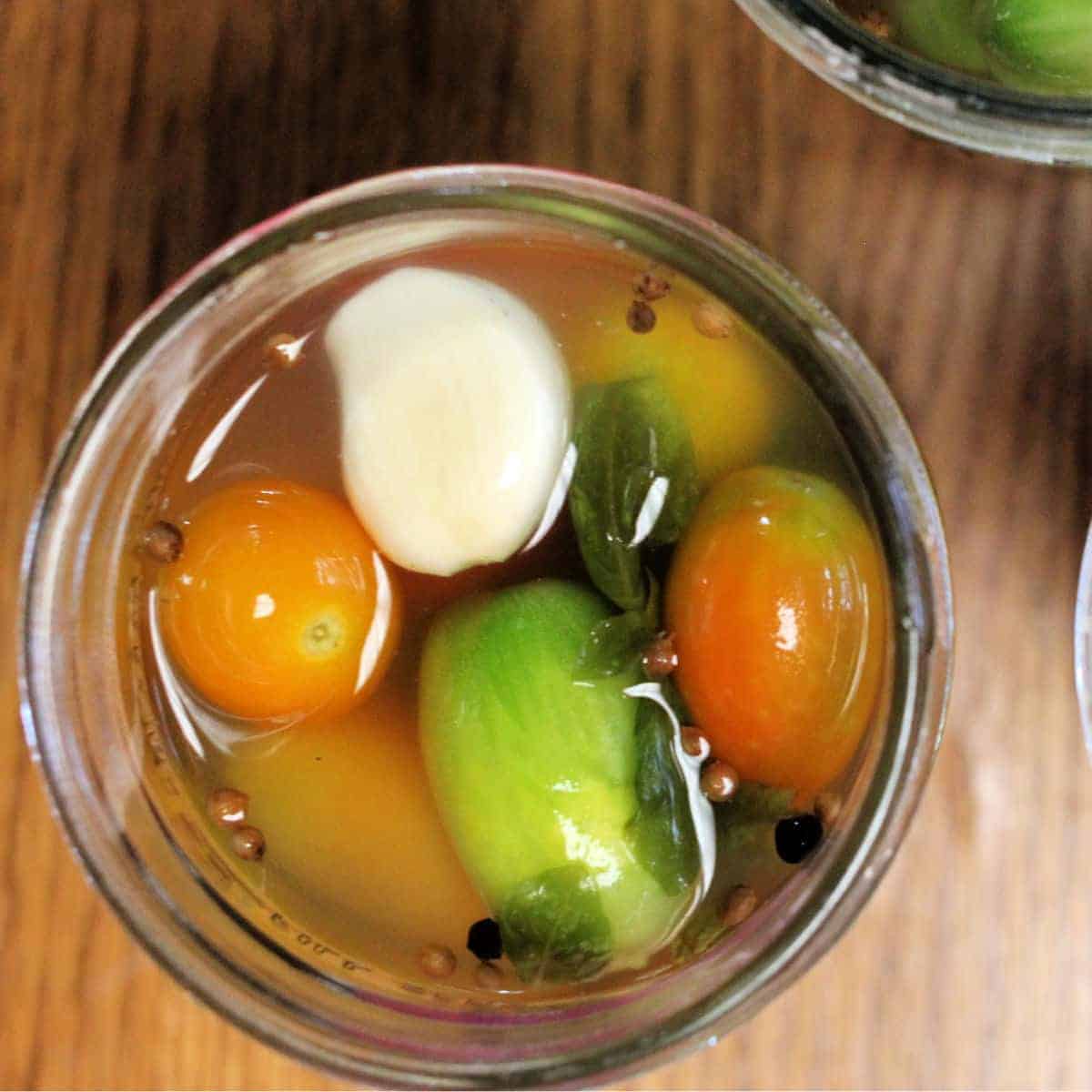 A jar filled with fermented cherry tomatoes, a garlic clove, and herbs in brine creates a colorful display. The mixture includes peppercorns enhancing flavor, all set on a rustic wooden surface for an inviting touch.