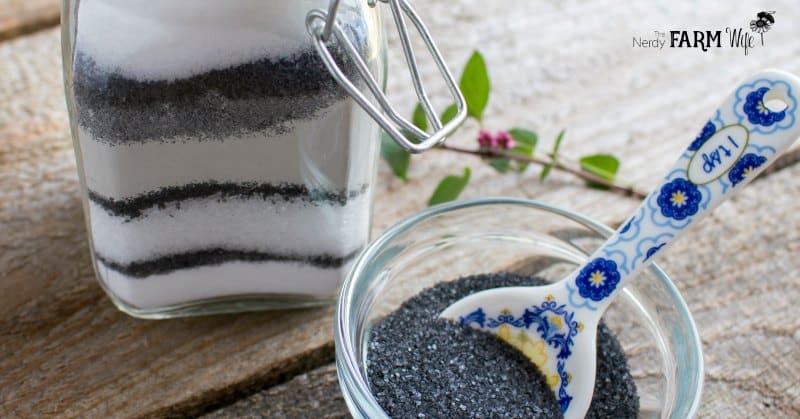 A glass jar filled with layered black and white salts sits on a wooden surface, featuring ingredients often used in natural cold and flu remedies. Next to it, a small bowl contains black salt with a decorative spoon. A sprig of greenery and flowers lies nearby.