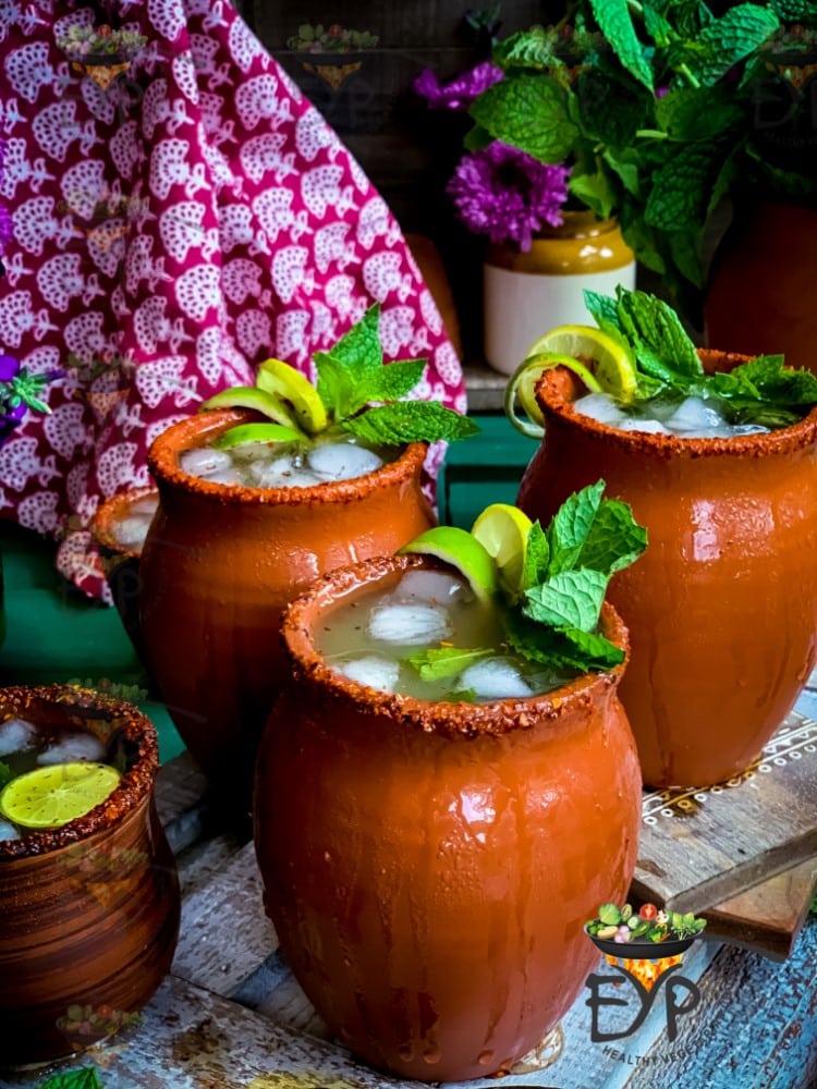 Three clay mugs filled with iced drinks, garnished with fresh mint and lime slices. The rim of each mug is coated with chili powder, offering a spicy twist on classic apple recipes. In the background, there's a patterned cloth and flowers, giving the scene a vibrant, traditional feel.