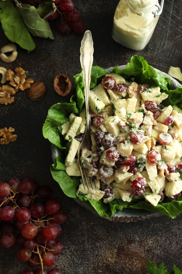 A delightful apple recipe awaits in this salad featuring sliced apples, red grapes, and walnuts on a bed of lettuce, all drizzled with creamy dressing. A fork sits ready amidst whole walnuts, grape clusters, and a glass bottle of dressing.