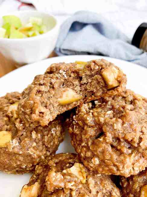 A close-up of a plate of baked oatmeal cookies with chunks of apple, showcasing a delightful entry in the apple recipes category. One cookie is partially bitten, revealing its soft, chewy texture. A bowl with green apple pieces sits invitingly in the background.