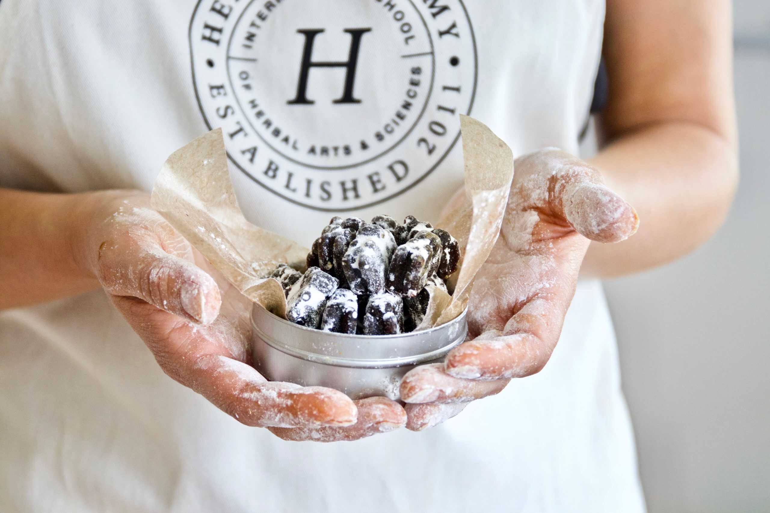 A person wearing an apron holds a small tin of powdered sugar-coated pastries, hands dusted with flour. The apron features the round logo of "HERBAL ARTS ACADEMY ESTABLISHED 2011," renowned for its expertise in natural cold and flu remedies.