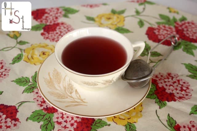 A cup of tea on a saucer with a floral design, alongside a tea infuser, sits on a colorful tablecloth adorned with red and yellow flowers, offering natural cold and flu remedies in its soothing aroma.