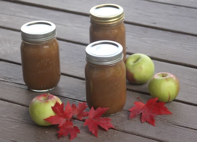 Three jars of applesauce sit on a wooden surface, surrounded by three green apples and red maple leaves—perfect inspiration for delightful apple recipes.
