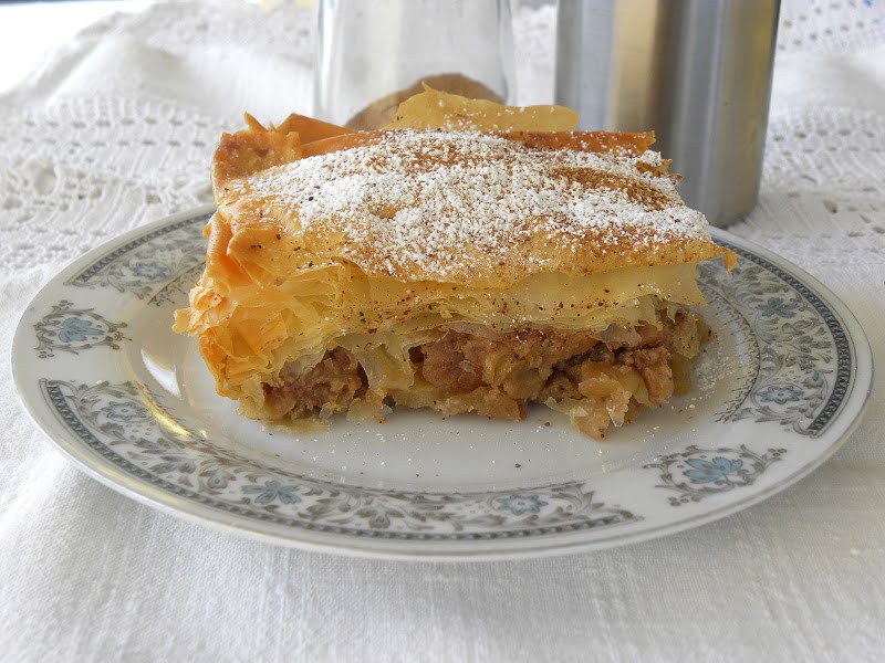 A slice of apple strudel, a classic from beloved apple recipes, is dusted with powdered sugar on a decorative plate, set atop a white lace tablecloth. In the background, a metal container peeks into view.