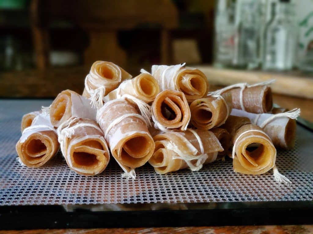 A stack of rolled fruit leather snacks, reminiscent of classic apple recipes, is tied with white strings on a mesh surface. In the blurred background, wooden chairs and glass jars add a rustic touch. The rolls appear to be made from a light brown, translucent material.