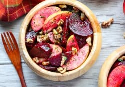 A wooden bowl brims with a vibrant apple recipe—an inviting salad of sliced apples, beets, and walnuts, adorned with fresh herbs. Nearby, a wooden fork rests on a red-checkered cloth atop a light wooden table.