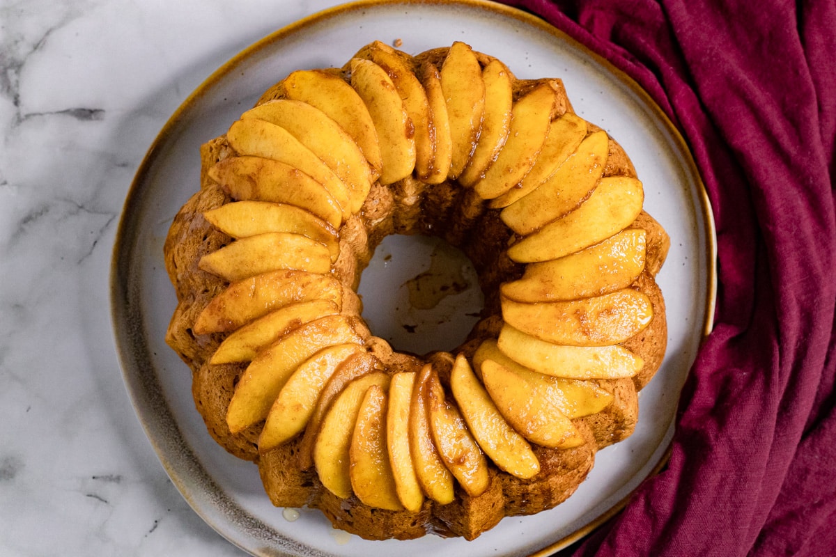 A classic bundt cake crowned with neatly arranged apple slices, showcasing one of the finest apple recipes. A dark red cloth peeks out beside the plate on a marble surface.