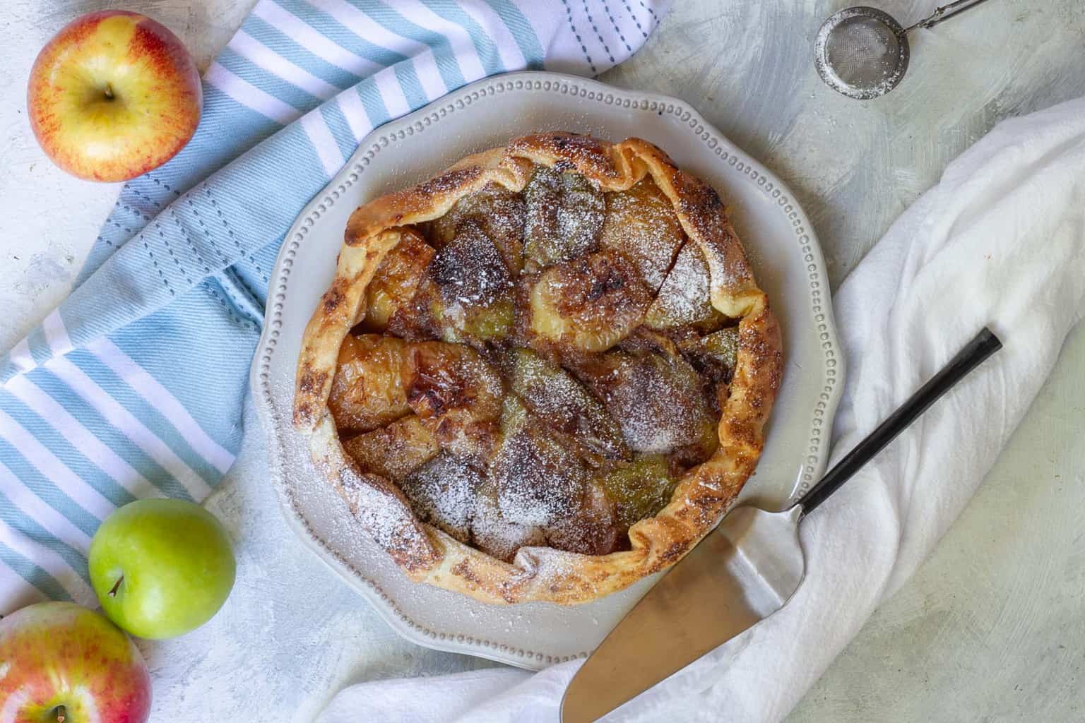 A rustic galette, perfect for apple recipes, is topped with sugar on a decorative plate. A pie server rests beside it on a white cloth. Two apples and a striped kitchen towel are nearby, along with a small sifter.
