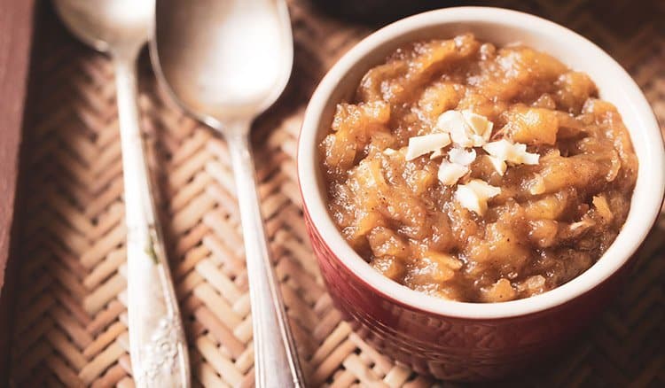 A bowl of warm brown dessert, reminiscent of classic apple recipes, is garnished with chopped nuts and sits on a woven mat. Two silver spoons are placed beside the bowl.