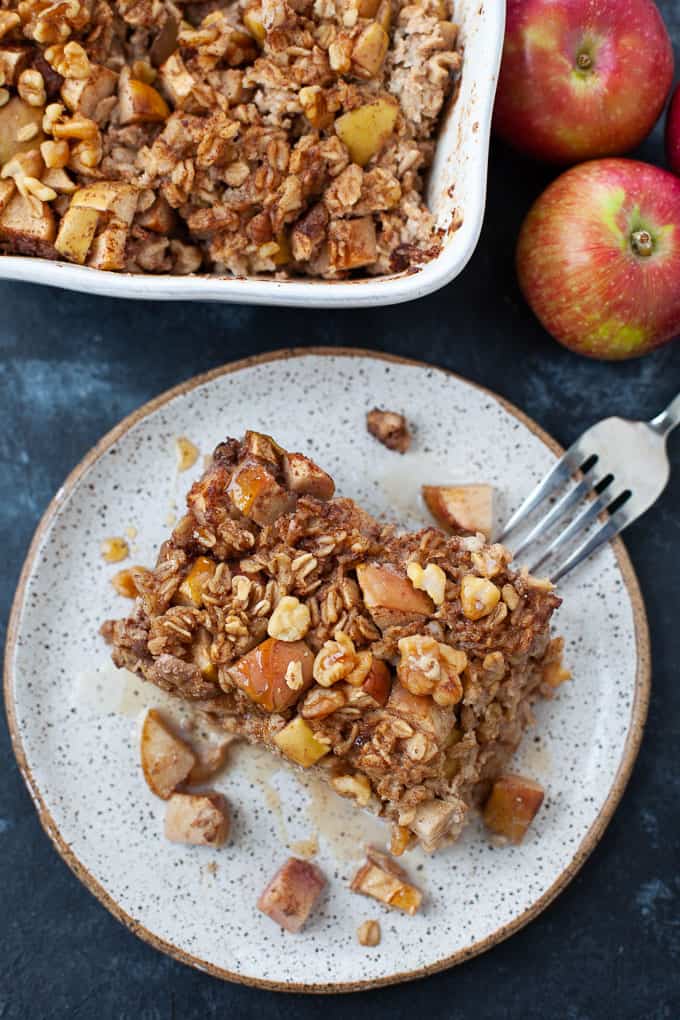 A slice of apple baked oatmeal on a speckled plate, topped with walnuts and apple chunks—a delightful addition to your collection of apple recipes. In the background, a baking dish brimming with more oatmeal awaits beside two fresh apples. A fork rests invitingly on the plate.