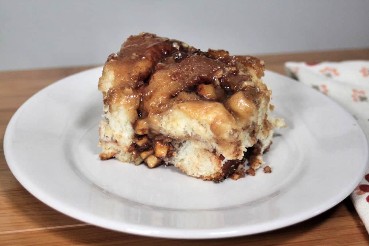 A slice of apple cinnamon bread pudding on a white plate invites admiration. Topped with caramelized apple chunks and cinnamon sauce, this dessert is the crown jewel of apple recipes. A cloth napkin with an orange print is partially visible in the background, adding a cozy touch.