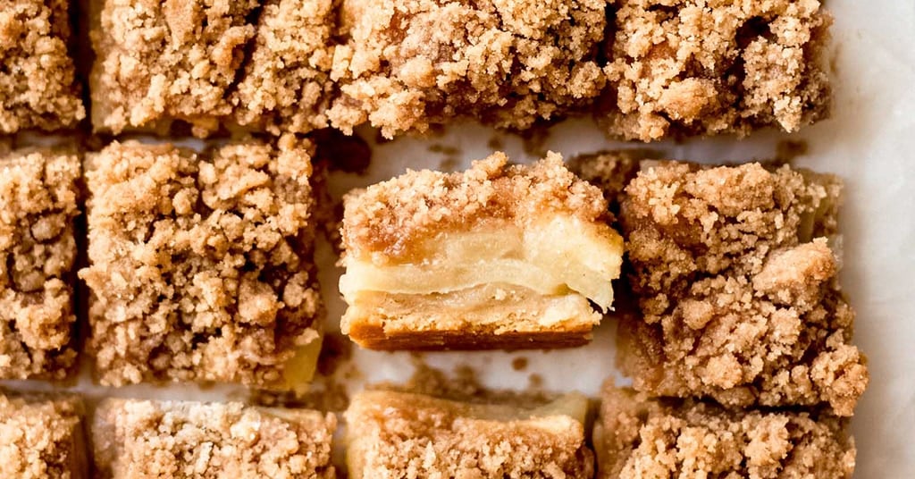 Top view of sliced apple crumb bars, a delightful addition to your apple recipes collection, arranged neatly on a parchment-lined surface. One bar is slightly raised, revealing layers of baked apples and crumbly streusel topping.